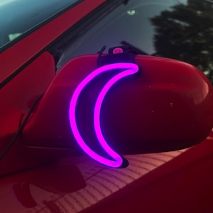 May include: A pink neon crescent moon shaped light attached to a red car's side mirror.