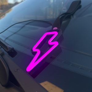 May include: A pink lightning bolt shaped car charm hanging from a black strap on a car windshield.