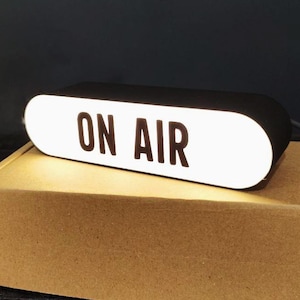 Peut inclure: Un panneau rectangulaire noir "ON AIR" avec des lettres blanches, éclairé sur un fond sombre. Le panneau est posé sur une boîte en carton marron. Le panneau est allumé, indiquant qu'il est en service.