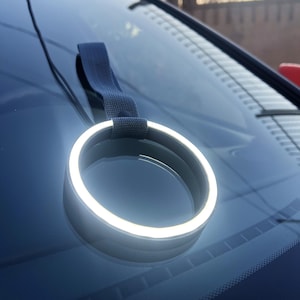 Puede incluir: Un anillo LED blanco con una correa negra unida a la parte superior. El anillo está colgado del parabrisas de un coche.