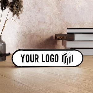 May include: A black and white rectangular sign with rounded edges, featuring the text "YOUR LOGO" in bold, black letters. A geometric logo is on the right. The sign is on a wooden surface.