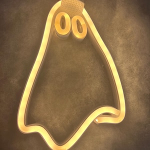 May include: A white neon light shaped like a ghost with two round eyes. The light is on and glowing yellow.