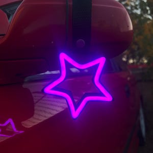 May include: A purple neon star-shaped car charm hanging from a black strap. The charm is attached to a red car's rearview mirror.