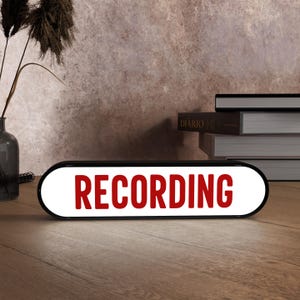 May include: A black and white illuminated sign with the word "RECORDING" in red letters. The sign is on a wooden surface, with a vase of dried plants and books in the background.