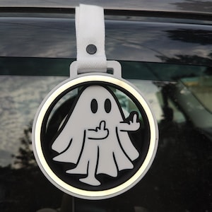 Op de afbeelding: Een wit en zwart spookvormig auto-sieraad met een middelvingergebaar. De charme is rond met een witte rand en een witte band aan de bovenkant. De charme hangt aan een autoruit.