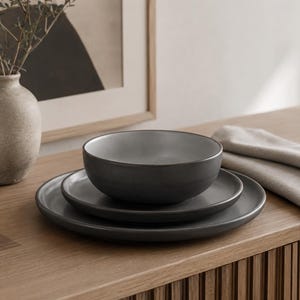 Op de afbeelding: Een set donkergrijs keramisch servies bestaande uit een groot dinerbord, een kleiner bord en een diepe kom die op elkaar zijn gestapeld. De stukken hebben een gladde, matte afwerking met een consistente donkergrijze tint over de hele set.