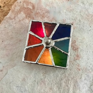 May include: A stained glass piece with a rainbow colour scheme. The glass is set in a silver frame and has a silver knob in the centre.