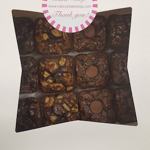May include: A white box with a clear window showing a variety of chocolate brownies. The brownies are arranged in a grid pattern and some have nuts and chocolate chips. The box has a pink and white sticker with the text "Nancy's Bake Shop" and "Thank you!"