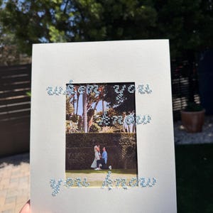 May include: A white rectangular card featuring a central rectangular cutout displaying a photograph of a proposal in a garden. The text "when you know you know" is embroidered in light blue thread around the cutout on the white card surface.