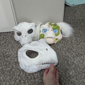 以下が含まれることがあります： 3つの恐竜マスクが写っています。2つは白で、1つは骸骨のようなデザイン、もう1つは詳細な顔です。3つ目のマスクは緑と白のアクセント、青い目、白い毛皮が付いています。