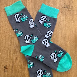 May include: A pair of gray socks with a teal green cuff. The socks have a repeating pattern of white skulls with teal green hearts and teal green boxes with white dots.