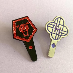 Op de afbeelding: Twee emaille pins. Eén is zwart met een rode glitter zeshoek en een leeuwenontwerp. De andere is een zilverkleurige pin met een klavervorm en een paars "H"-ontwerp.
