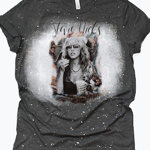 Puede incluir: Una camiseta gris oscura con un diseño de salpicaduras blancas que presenta una foto en blanco y negro de Stevie Nicks. El texto "Stevie Nicks" está escrito en cursiva negra sobre la foto.