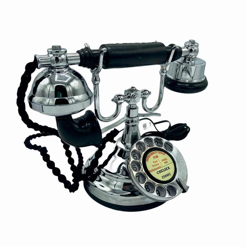 Pode incluir: Um telefone rotativo de estilo vintage com um auricular preto e uma base cromada. O telefone tem um dial preto com n&uacute;meros brancos e o texto "EMERGENCY CALLS FOR Fire - Dial 999 Police - Dial 999 Ambulances - Dial 999 CHELSEA 252497".