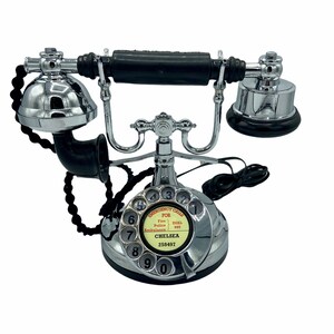 Pode incluir: Um telefone rotativo de estilo vintage com um auricular preto e uma base cromada. O telefone tem um dial branco com n&uacute;meros pretos e um r&oacute;tulo vermelho que diz "Chamadas de emerg&ecirc;ncia Para Bombeiros Discar 999 Pol&iacute;cia 999 Ambul&acirc;ncia 999 Chelsea 258497".