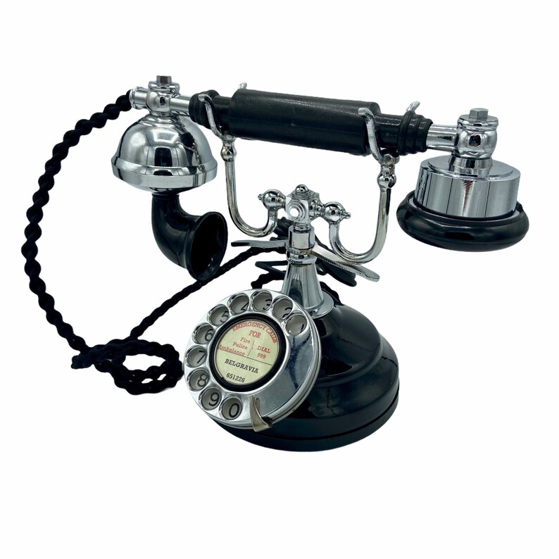 Pode incluir: Um telefone de disco preto com acabamento cromado. O telefone tem um cabo preto e uma base preta. O dial tem n&uacute;meros brancos e as palavras "P.O.B.  Push  Pulse  Exchange  999  BELGRAVIA  681228" impressas nele.