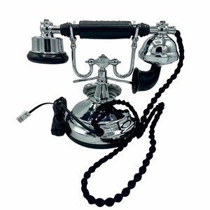 Pode incluir: Um telefone rotativo de estilo vintage com um auscultador preto e uma base cromada. O telefone tem um cabo preto e um cabo tran&ccedil;ado preto.