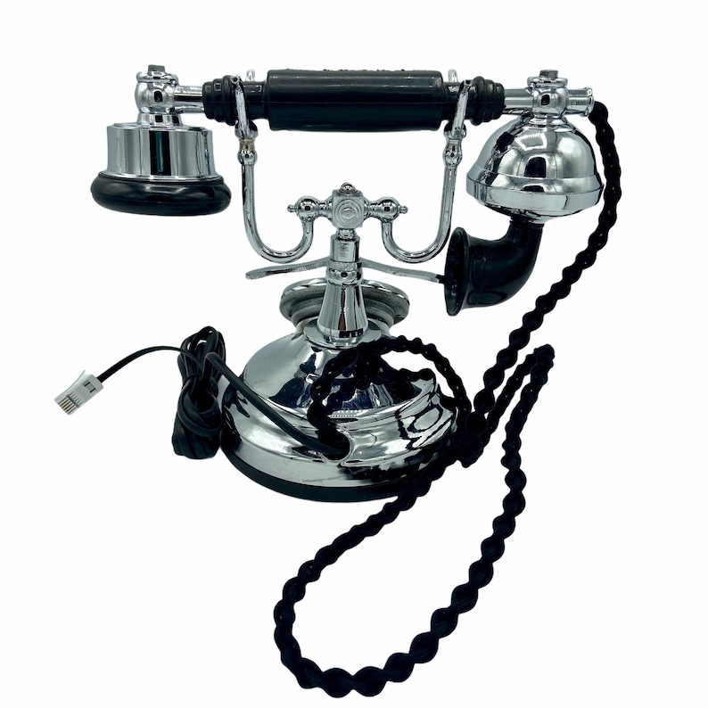 Pode incluir: Um telefone rotativo de estilo vintage com um auscultador preto e uma base cromada. O telefone tem um cabo preto e um cabo tran&ccedil;ado preto.