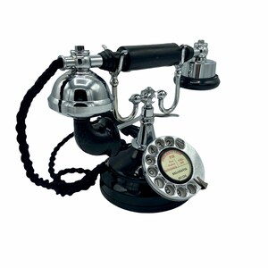 Pode incluir: Um telefone de disco preto com acabamento cromado. O telefone tem um cabo preto e um dial branco com n&uacute;meros pretos. O dial tem um r&oacute;tulo vermelho que diz "Chamadas de emerg&ecirc;ncia" e "Para bombeiros, pol&iacute;cia, ambul&acirc;ncia, disque 999". O r&oacute;tulo tamb&eacute;m diz "Belgravia 661 229".