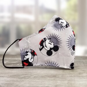 Puede incluir: Una mascarilla gris con un patrón repetido de Mickey Mouse en un diseño circular con rayos de sol. La mascarilla tiene presillas negras para las orejas y está hecha de tela. El fondo es una vista borrosa de una ventana.