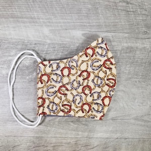 Puede incluir: Una mascarilla facial beige con una banda elástica blanca. La mascarilla está cubierta con un patrón de herraduras rojas, amarillas y moradas.