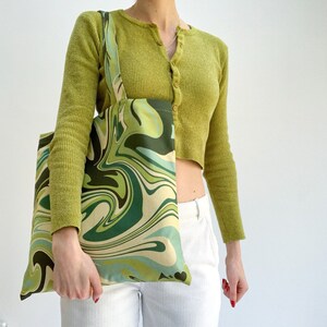 May include: A green and cream-colored tote bag with a swirling, marbled pattern. The bag has a rectangular shape with matching straps. The design incorporates shades of green and cream. The bag is held by a person.