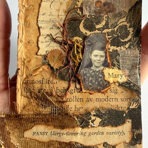 May include: A mixed media collage card featuring a black and white photograph of a child named Mary, layered over vintage paper with German text, floral patterns, torn brown paper edges, loose brown and yellow threads, and a label reading PANSY.