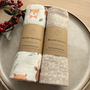 Könnte beinhalten: Zwei gerollte Musselintücher mit braunen Papieretiketten. Eines zeigt ein Fuchs- und Blumenmuster, das andere ein Blumenmuster. Die Etiketten lauten "Musselin Spucktuch 100% Baumwolle RatzFratzHandmade find us @ etsy & instagram."