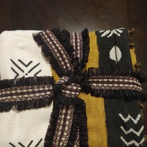 May include: A white, black, and gold woven throw blanket with black and brown ribbon tied in a bow. The blanket has a geometric pattern and is decorated with black fringe.