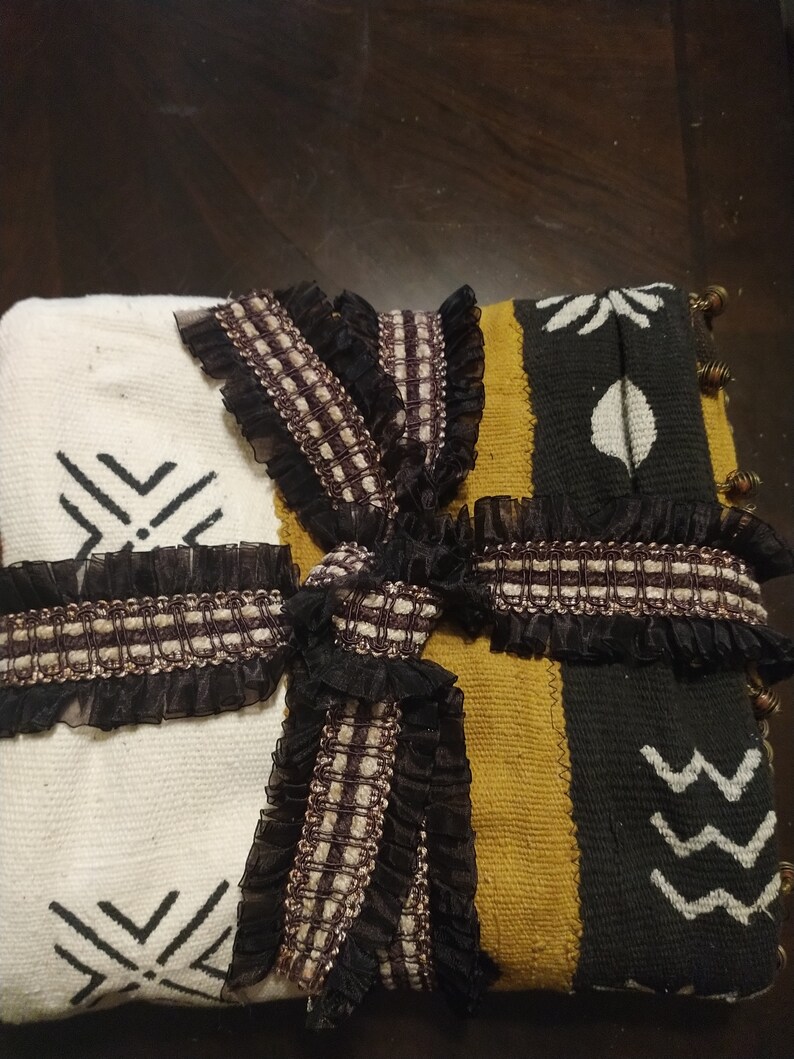 May include: A white, black, and gold woven throw blanket with black and brown ribbon tied in a bow. The blanket has a geometric pattern and is decorated with black fringe.