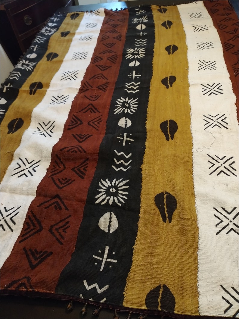 May include: A woven tapestry with a repeating geometric pattern in black, white, brown, and gold. The tapestry has a fringe border.