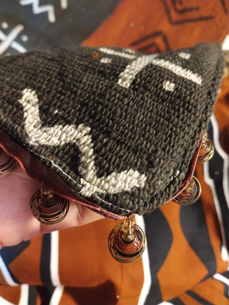 May include: A black and white patterned fabric pillow with a burgundy trim and brown beaded fringe.