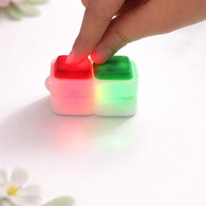 May include: A small, white, dual-button device with red and green illuminated squares. The device is being pressed by a finger. The buttons glow, suggesting an electronic function. The overall aesthetic is modern and minimalist.