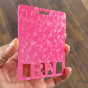 May include: A bright pink badge holder with a geometric pattern. The letters "RN" are cut out at the bottom. The holder has a rectangular shape with a slot at the top for a lanyard or clip.