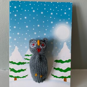 May include: A gray knitted owl with white eyes, a red heart, and yellow accents is displayed on a card with a winter scene. The card features a blue sky with white stars, a white moon, and two green trees covered in snow.
