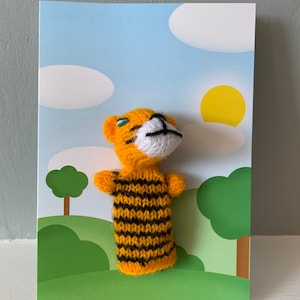 May include: A yellow and black striped knitted finger puppet of a tiger with green eyes and a white nose and mouth. The puppet is on a card with a blue sky, white clouds, a yellow sun, and green trees.