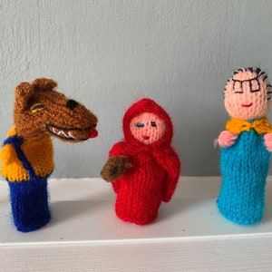 May include: Three knitted finger puppets: a brown wolf with a yellow shirt and blue pants, a red hooded figure with a brown basket, and a blue figure with a yellow scarf and glasses.