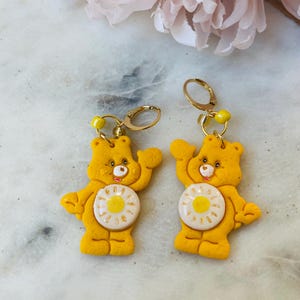 May include: A pair of yellow Care Bear earrings with a white daisy on the front. The earrings are made of polymer clay and have gold-toned hooks.
