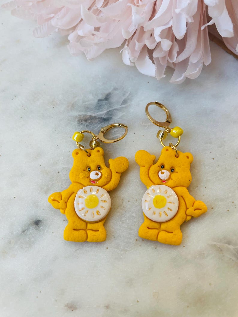 May include: A pair of yellow Care Bear earrings with a white daisy on the front. The earrings are made of polymer clay and have gold-toned hooks.