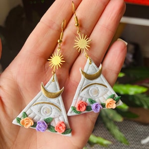 May include: A pair of white triangle earrings with a gold crescent moon, a gold star, and a glittery eye design. The earrings are decorated with small pink, orange, and purple flowers.