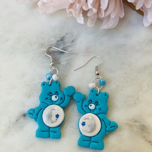 May include: A pair of blue Care Bear earrings with a moon and stars design. The earrings are made of polymer clay and have silver hooks.