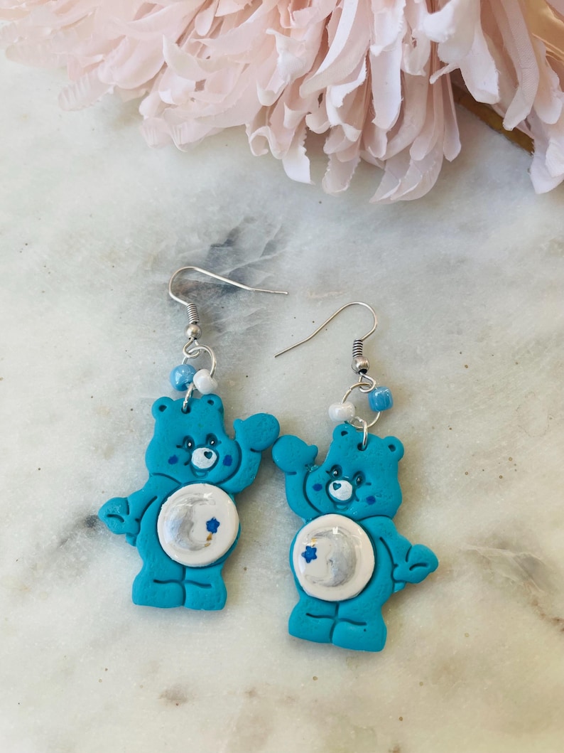 May include: A pair of blue Care Bear earrings with a moon and stars design. The earrings are made of polymer clay and have silver hooks.