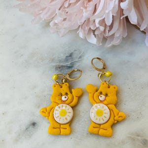 May include: A pair of yellow Care Bear earrings with a white daisy on the front. The earrings are made of polymer clay and have gold hoops.