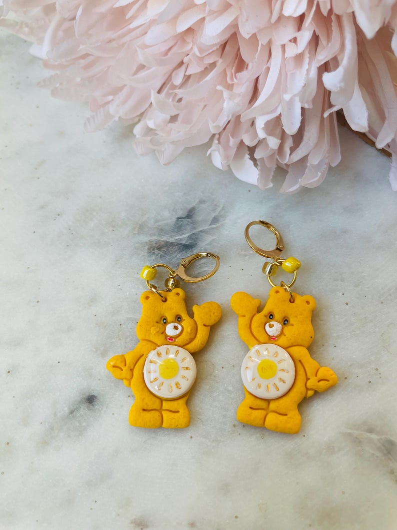 May include: A pair of yellow Care Bear earrings with a white daisy on the front. The earrings are made of polymer clay and have gold hoops.