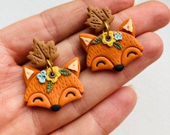 May include: A pair of orange fox-shaped earrings. Each earring features a fox face with a black nose, a floral crown, and a brown leaf accent. The earrings are connected by a gold-colored wire.