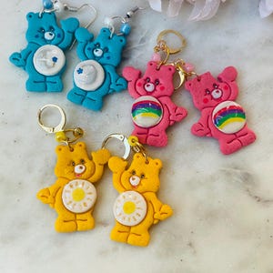 May include: A set of six Care Bear earrings. Two pairs are blue with a star, two pairs are pink with a rainbow, and two pairs are yellow with a sun.