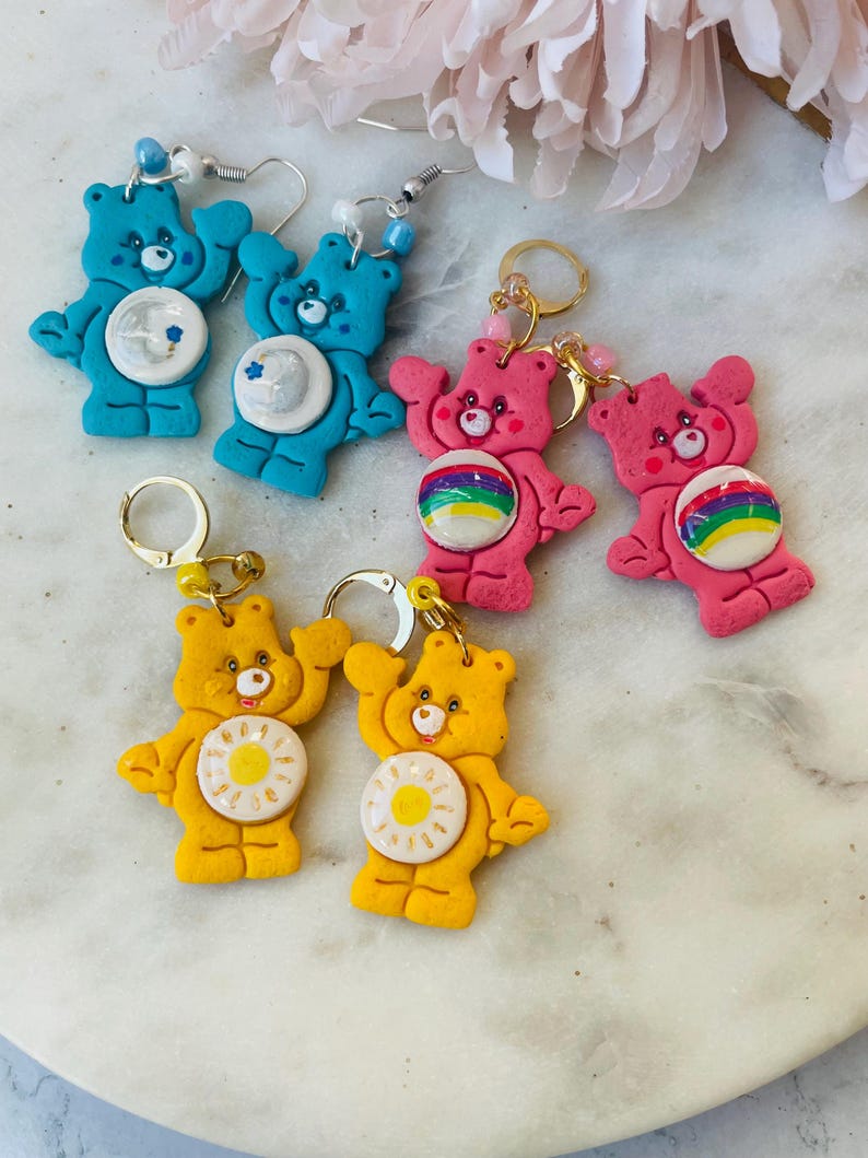 May include: A set of six Care Bear earrings. Two pairs are blue with a star, two pairs are pink with a rainbow, and two pairs are yellow with a sun.