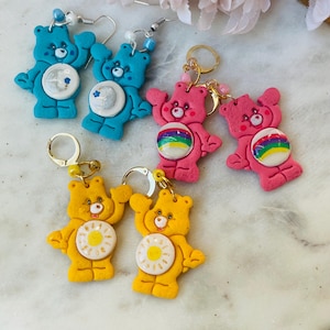 May include: A set of six Care Bear earrings. The earrings are made of polymer clay and feature different Care Bear characters. The earrings are available in blue, pink, and yellow.