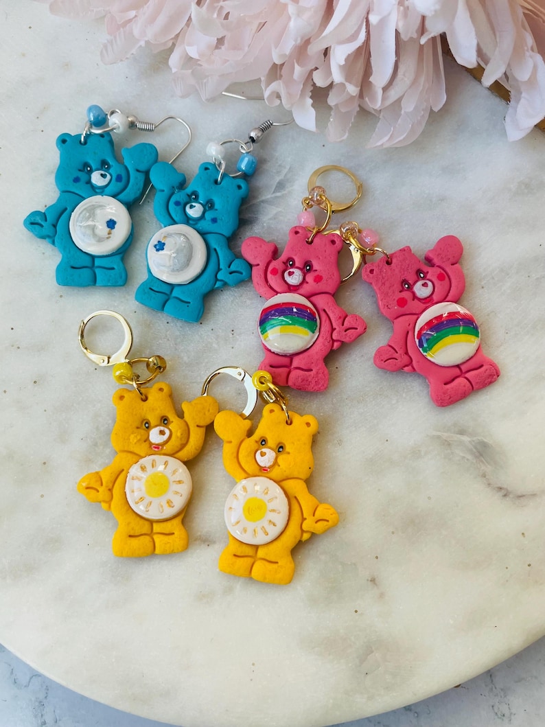 May include: A set of six Care Bear earrings. The earrings are made of polymer clay and feature different Care Bear characters. The earrings are available in blue, pink, and yellow.