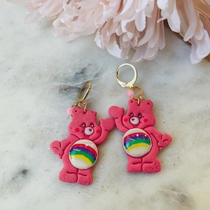 May include: A pair of pink Care Bear earrings with rainbow belly buttons. The earrings are made of polymer clay and have gold hoops.